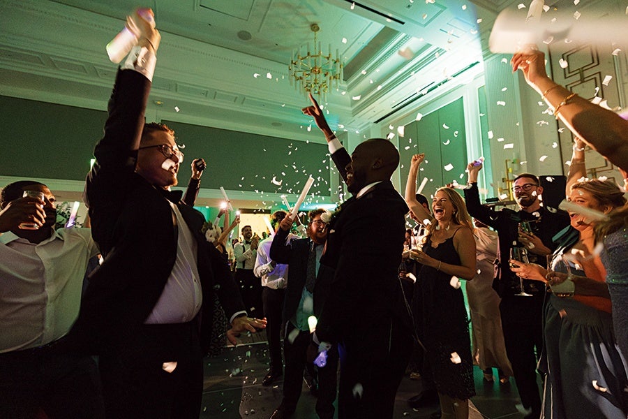 A lively indoor celebration with a group of people dressed in formal attire, dancing and cheering. Confetti is falling from above, and several individuals are holding drinks and light sticks. The room features elegant decor with a chandelier and soft green lighting.