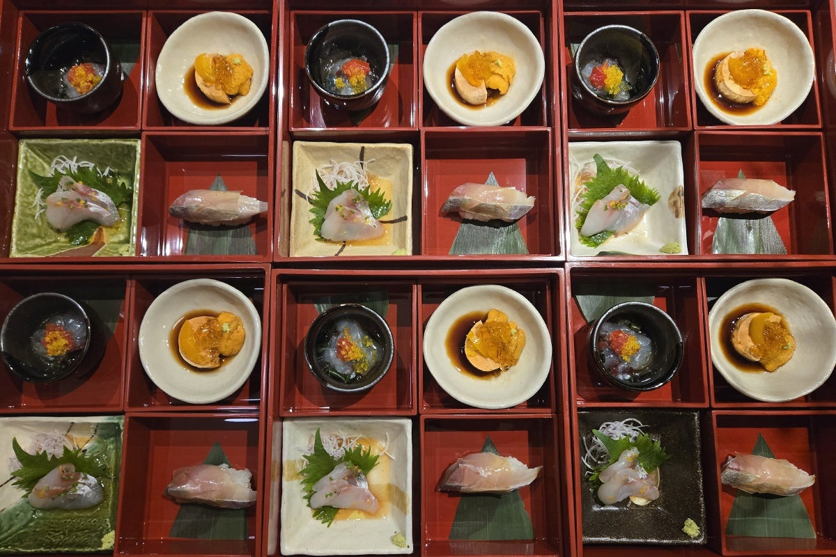 A neatly arranged Japanese bento box with multiple compartments, each containing small servings of sashimi on green shiso leaves, a piece of fish nigiri, and a small bowl with sea urchin (uni) in soy sauce. The presentation is symmetrical and visually appealing with a red lacquered tray background.