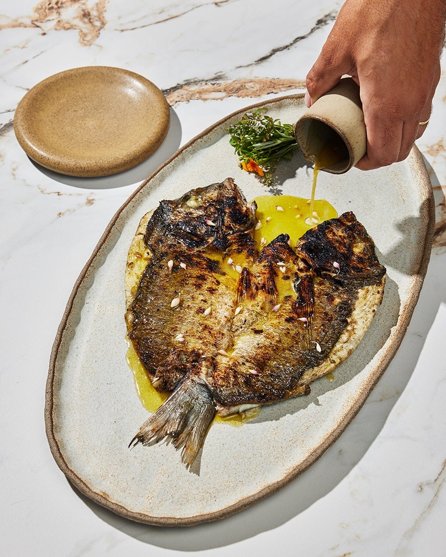 A hand pours olive oil onto a butterflied, whole grilled fish, garnished with a sprig of fresh herbs.