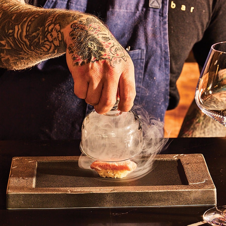 A tattooed hand is lifting a glass dome releasing smoke from a piece of sushi placed on a rectangular stone plate. The person is wearing a dark shirt and a blue apron with the word "bar" visible. A wine glass is partially visible on the right side.