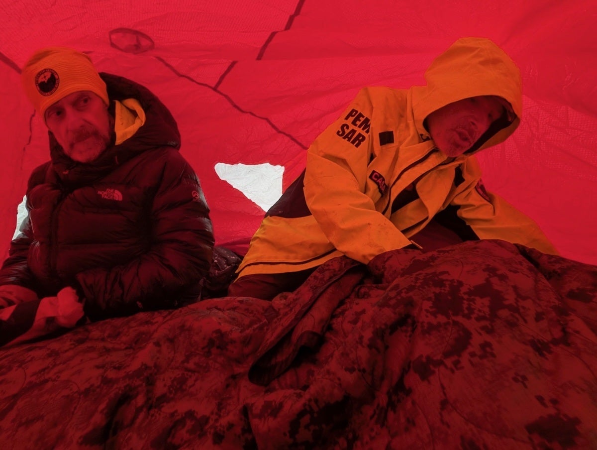 Two men inside a red tent, one wearing a black puffer jacket and yellow beanie, the other in a yellow hooded jacket with "PEMA SAR" written on the sleeve, attending to someone or something covered with a dark patterned blanket.