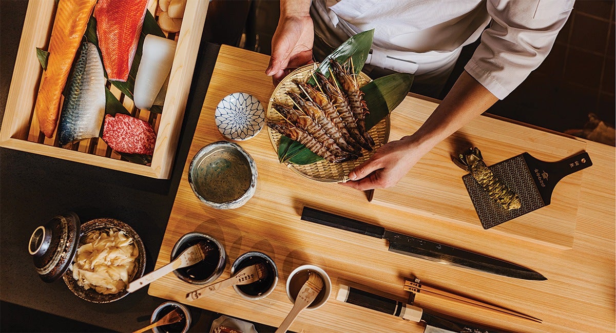 A wooden table set for sushi preparation with various ingredients and tools. A person in a white chef's coat holds a bamboo basket containing several large shrimp on green leaves. To the left, a wooden box contains assorted fish fillets and a piece of marbled meat. On the table, there are small bowls with sauces and brushes, a bowl of pickled ginger, a large knife, chopsticks, and a wasabi root on a grater.
