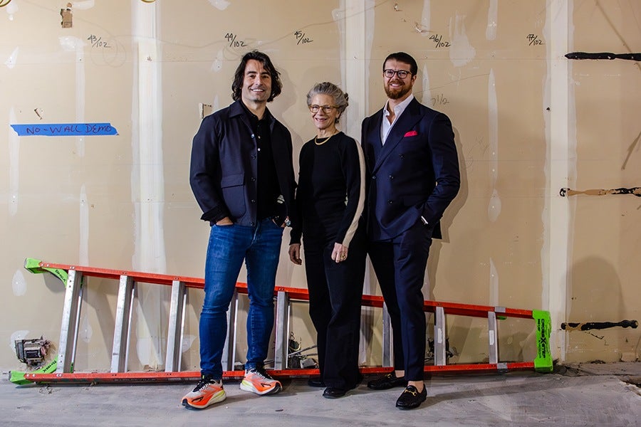 Three people stand indoors in front of an unfinished wall with markings and a red and silver ladder lying horizontally on the floor behind them. The person on the left wears a dark jacket, blue jeans, and colorful sneakers. The person in the middle wears a black outfit with beige stripes on the sleeves and glasses. The person on the right wears a dark suit with a white shirt and a red pocket square, along with black loafers.