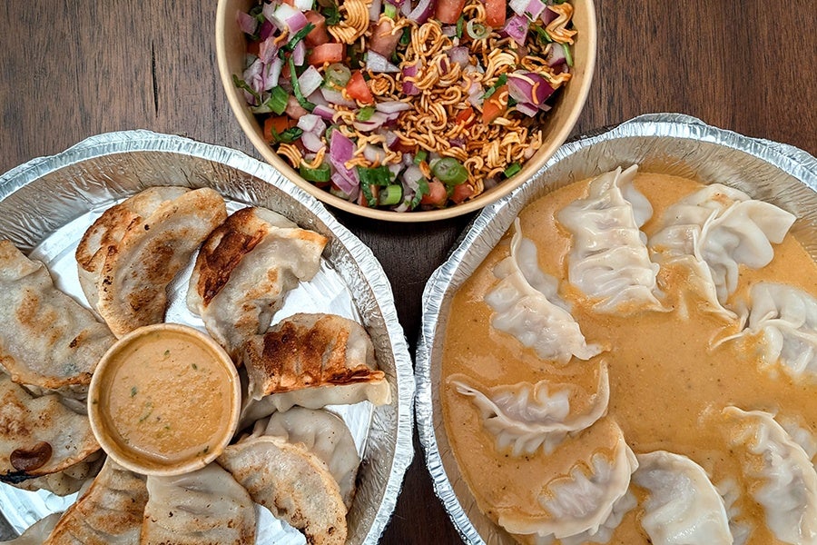 Two foil containers of dumplings are shown, one with pan-fried dumplings arranged around a small bowl of dipping sauce, and the other with steamed dumplings in a creamy orange sauce. Above them is a bowl of salad containing chopped onions, tomatoes, green chilies, and crispy noodles.