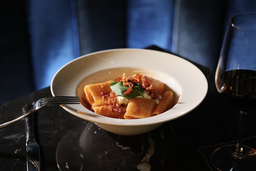 A white bowl contains large square pasta pieces topped with crispy bacon bits, a dollop of creamy sauce, and a green basil leaf. The bowl is placed on a dark marble table with a fork resting on the edge, a knife beside it, and a glass of red wine to the right. The background features a dark blue cushioned surface.