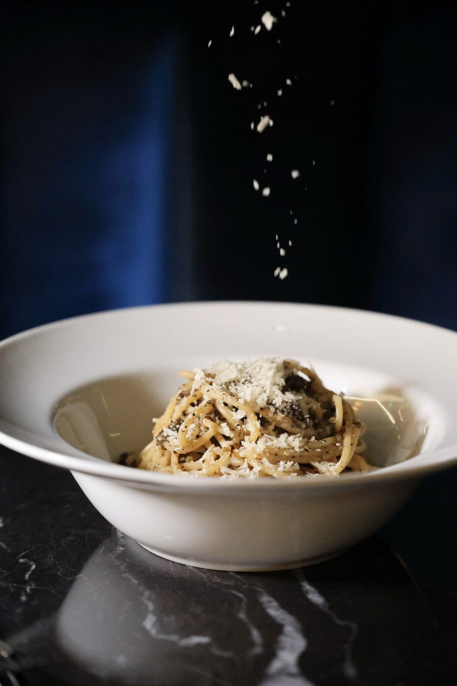 A white bowl filled with spaghetti topped with grated cheese and black pepper, with additional grated cheese falling onto the pasta. The bowl is placed on a dark marble surface.