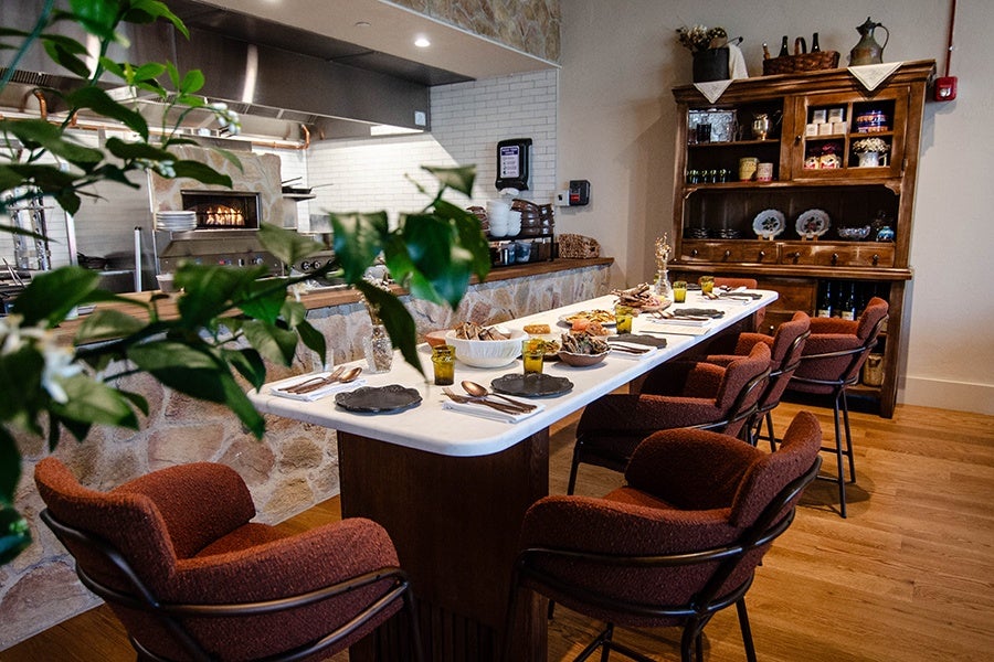 A long white table at a restaurant has six upholstered maroon chairs and looks into an open kitchen with a fire burning in an oven.