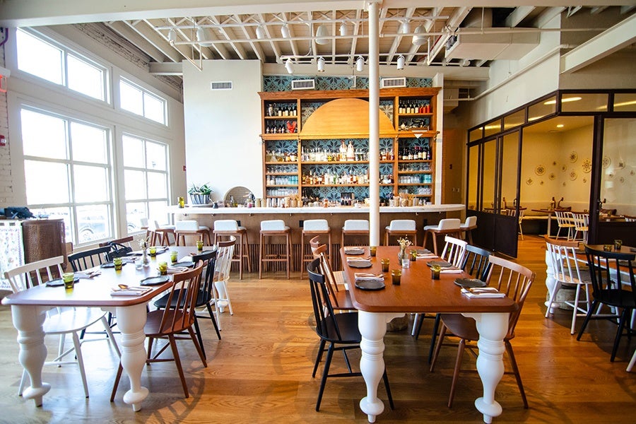 A brightly lit restaurant has a rustic, homey vibe, with wooden tables and chairs and Greek-style plates hanging on a wall.