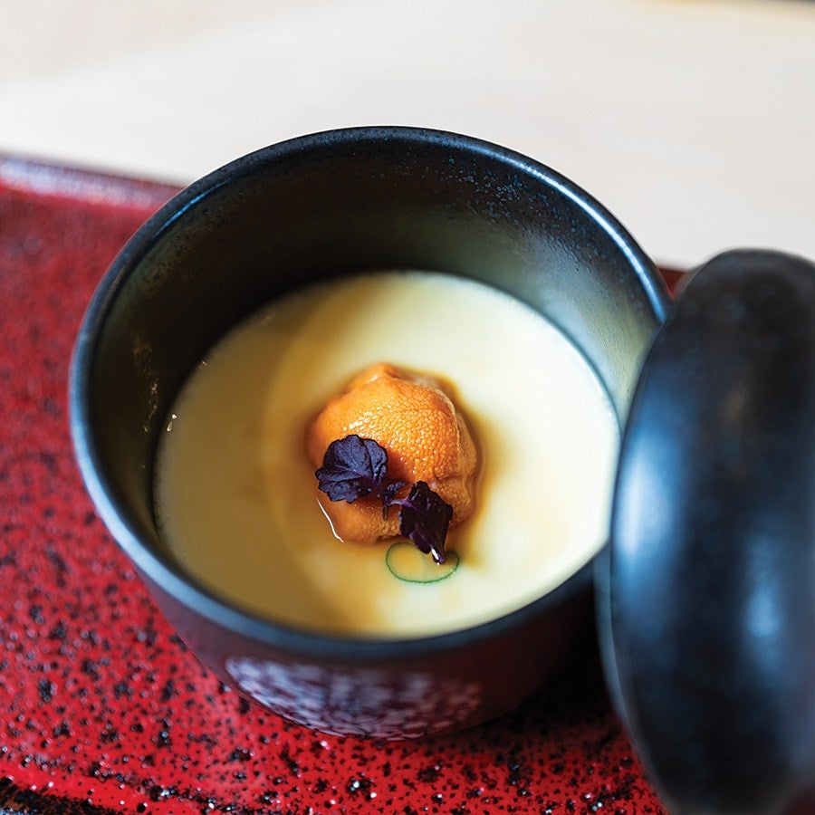 A black ceramic bowl with a partially open lid contains a creamy yellow custard topped with a piece of orange sea urchin and two small dark purple leaves. The bowl is placed on a red speckled tray.