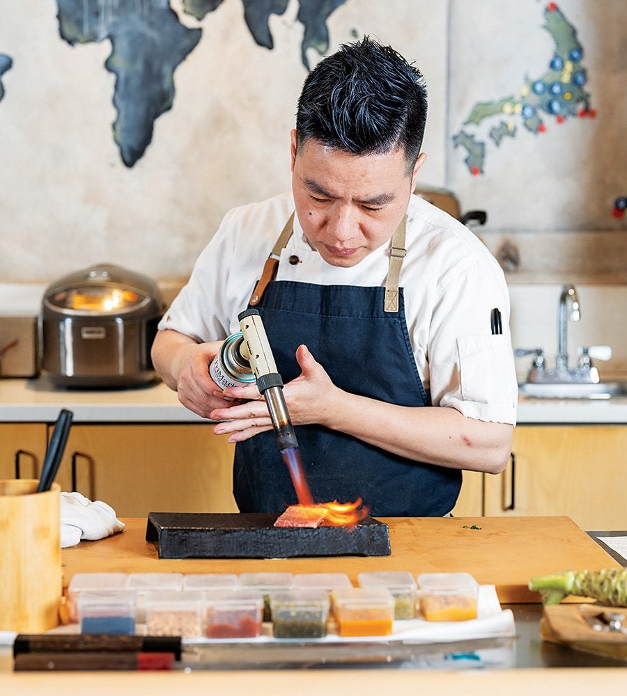 A chef wearing a white shirt and dark apron is using a blowtorch to sear a piece of meat on a black rectangular cooking surface. The setting appears to be a kitchen with a wooden countertop, various containers of spices or ingredients in the foreground, and a map on the wall in the background.