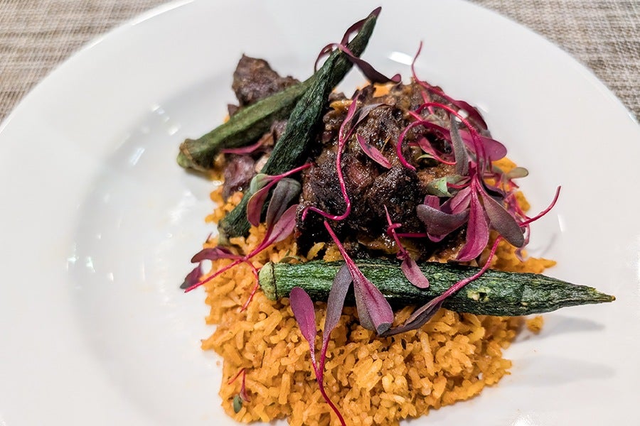 A dish featuring seasoned yellow rice topped with cooked okra, pieces of dark-colored meat, and garnished with purple microgreens, served on a white plate.