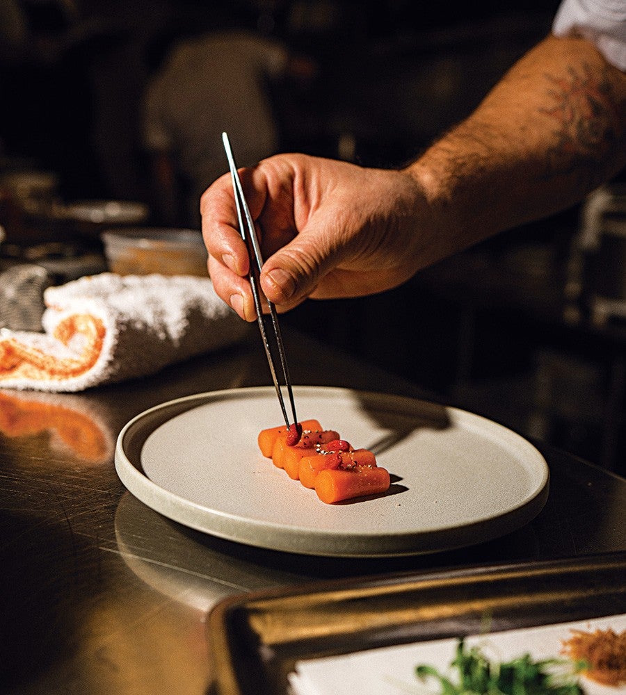 A hand with a tattoo on the forearm is using metal tweezers to place a small garnish on a row of six neatly arranged orange cylindrical food pieces on a plain, round, light-colored plate. The setting appears to be a kitchen with a towel and other kitchen items blurred in the background.