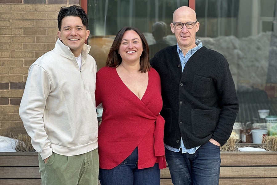 Three people stand side by side outdoors in front of a window and brick wall. The person on the left wears a light beige quarter-zip sweater and green pants. The person in the middle wears a red wrap-style top and dark jeans. The person on the right wears glasses, a black buttoned cardigan over a light blue shirt, and dark jeans. All three are smiling.