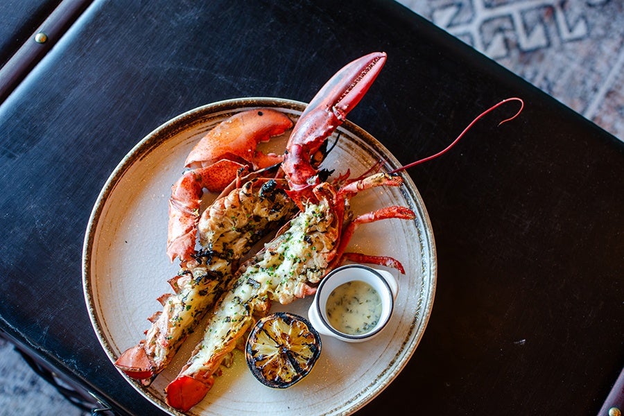 Grilled lobster split in half on a beige plate, garnished with herbs, accompanied by a charred lemon half and a small cup of creamy herb sauce, placed on a dark wooden surface.