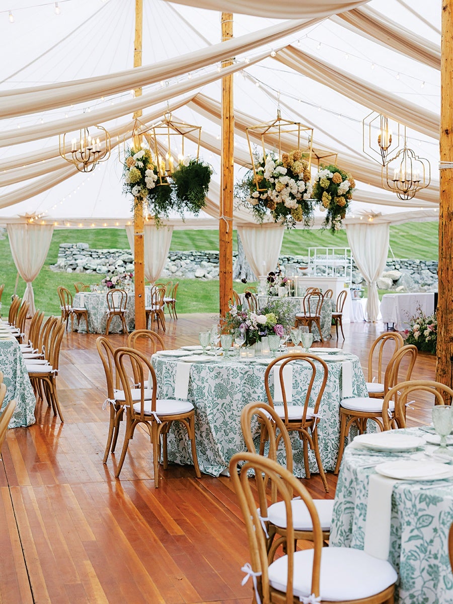 Outdoor wedding reception setup under a white tent with draped fabric and string lights. Round tables are covered with green and white floral tablecloths, each surrounded by wooden chairs with white cushions. Hanging gold geometric lanterns adorned with white and green floral arrangements are suspended from the tent ceiling. The floor is polished wood, and the background shows green grass and a stone wall.