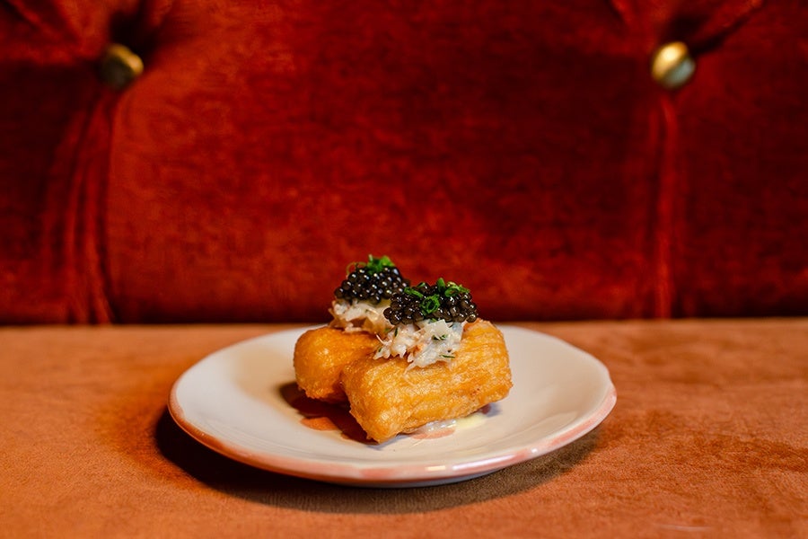 Two rectangular pieces of fried food topped with a creamy mixture and black caviar, garnished with chopped green herbs, served on a white plate against a red velvet background.