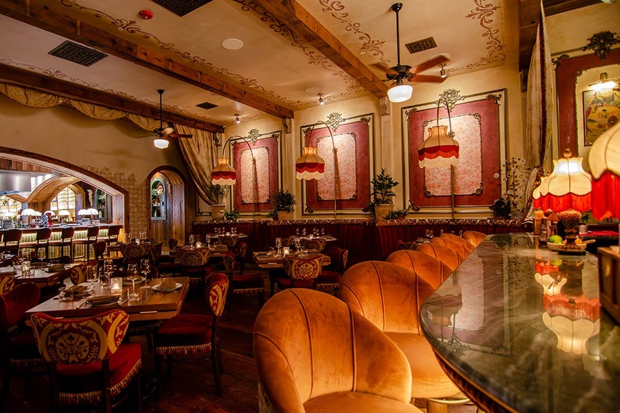 A warmly lit, vintage-style restaurant interior featuring ornate upholstered chairs with red and gold patterns, wooden tables set with glassware and plates, and a curved marble bar with plush orange velvet stools. The walls are decorated with large framed panels in red and cream tones, and hanging lamps with scalloped red and cream shades provide soft lighting. The ceiling has wooden beams and decorative stenciling, adding to the cozy, elegant atmosphere.
