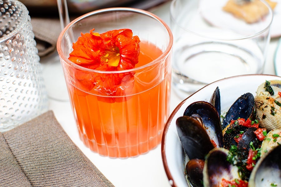 A clear glass filled with an orange-colored drink garnished with a bright orange edible flower, placed next to a bowl of mussels and clams topped with chopped herbs and red seasoning. A textured glass and a folded brown napkin are also visible nearby.