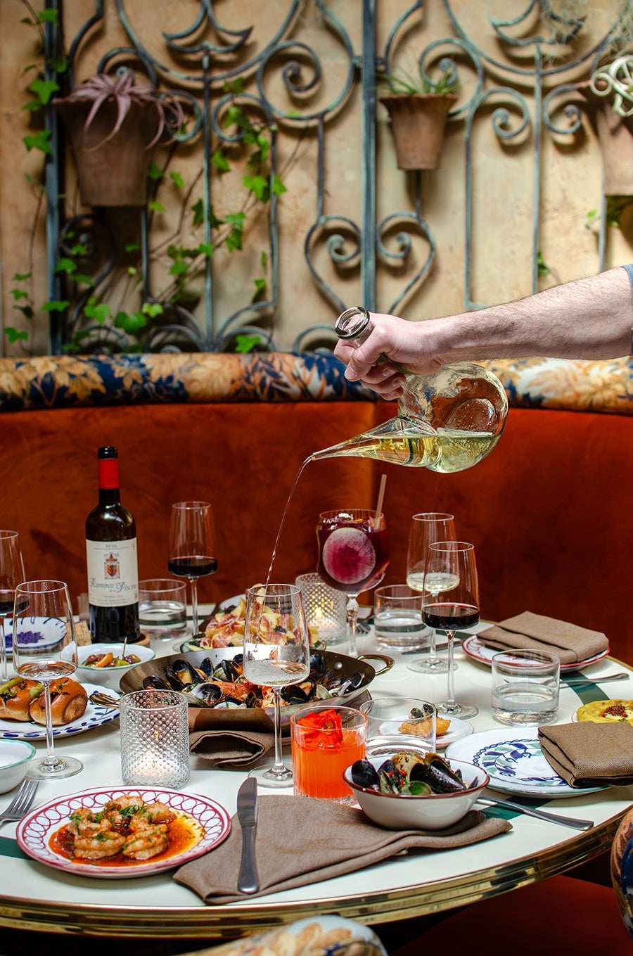 A table set with various dishes and drinks, including a plate of shrimp in sauce, a bowl of mussels, and a pan of seafood. Several wine glasses, a bottle of red wine, and a glass with an orange cocktail are also present. A hand is pouring white wine from a uniquely shaped glass decanter into a wine glass. The background features a cushioned bench with floral upholstery and a decorative wrought iron wall with potted plants.
