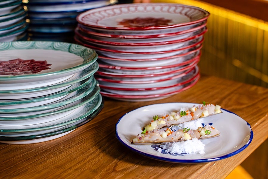 Two razor clam shells served on a white plate with a blue rim, topped with diced vegetables and herbs, accompanied by a small pile of coarse salt. In the background, there are stacks of decorative plates with green and red patterns on a wooden surface.