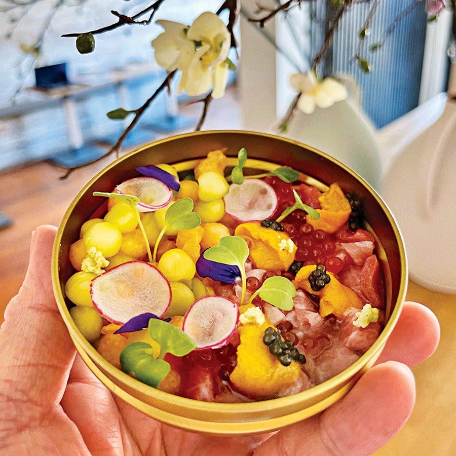 A hand holding a round, gold-colored tin filled with a colorful gourmet dish. The dish includes yellow spheres, thin radish slices, small green leaves, orange sea urchin pieces topped with black caviar, and translucent red fish roe, all arranged artfully. The background shows blurred indoor elements and white flowers on branches.