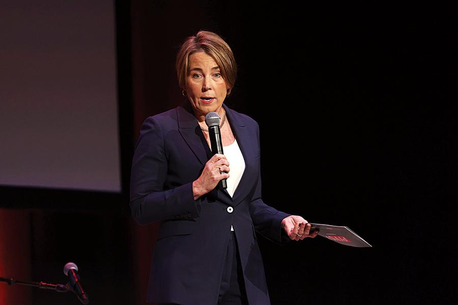A woman with short blonde hair wearing a dark navy blazer and white top is speaking into a handheld microphone. She is holding a piece of paper or card in her other hand and appears to be on stage with a dark background.