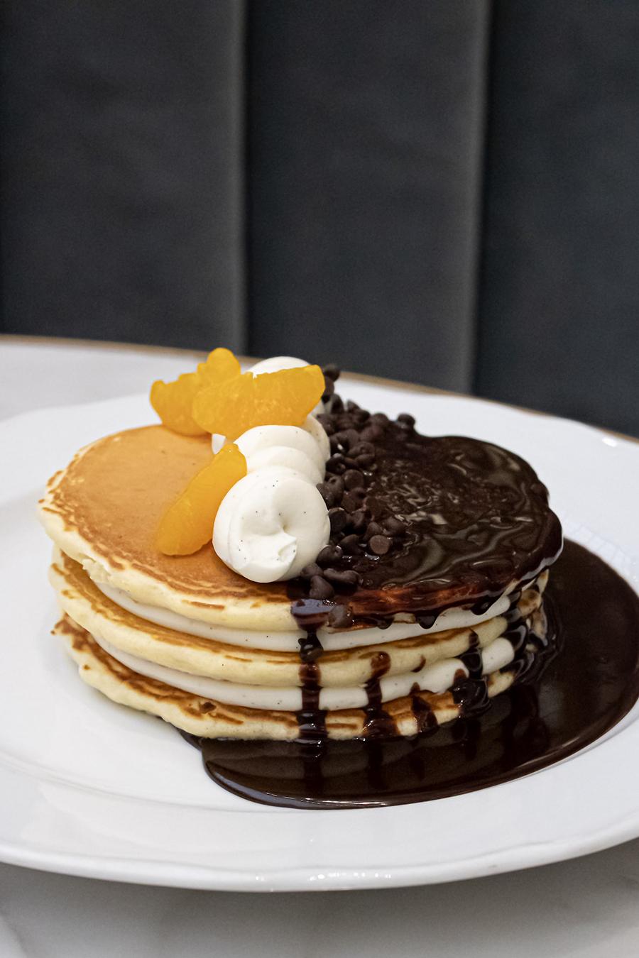 A stack of pancakes is topped with chocolate sauce, chocolate chips, a swirl of ricotta, and orange slices