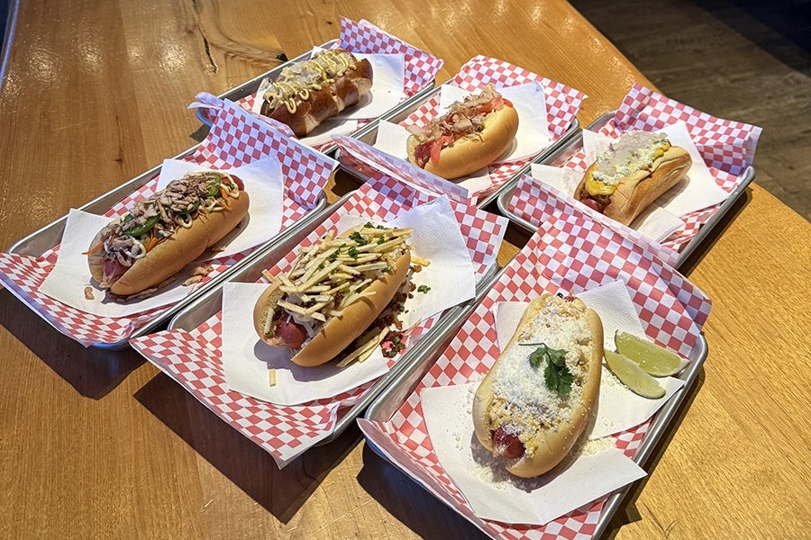 Six hot dogs are served on metal trays lined with red and white checkered paper. Each hot dog has different toppings, including shredded cheese, mustard, chopped herbs, potato sticks, and lime wedges. The trays are arranged on a wooden table.