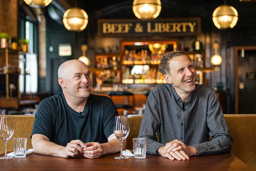 Two men sitting at a wooden table in a restaurant with a sign reading "BEEF & LIBERTY" in the background. The man on the left is bald and wearing a black t-shirt, while the man on the right has short hair and is wearing a gray button-up shirt. Both are smiling and looking slightly to the right. The table has empty wine glasses and water glasses in front of them. The restaurant interior features warm lighting and a cozy atmosphere.