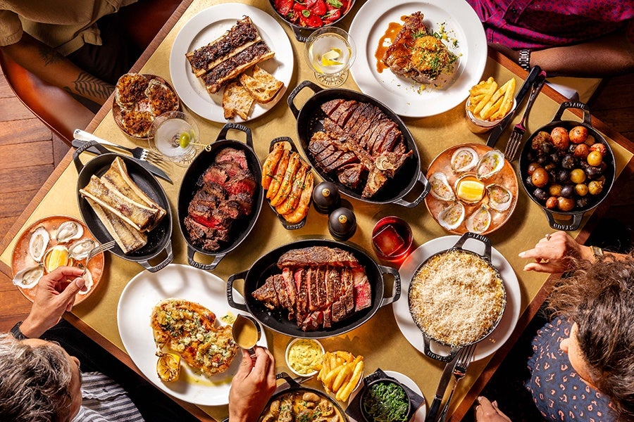 A table set with a variety of dishes including several plates of sliced steak in cast iron skillets, roasted potatoes, French fries, oysters on the half shell with lemon, grilled shrimp, a baked dish topped with melted cheese, bone marrow, a plate with two stuffed or topped pieces of bread, and a plate with a cooked chicken thigh. There are also drinks, including a glass with a lemon slice and a red beverage. Four people are seated around the table, reaching for food.