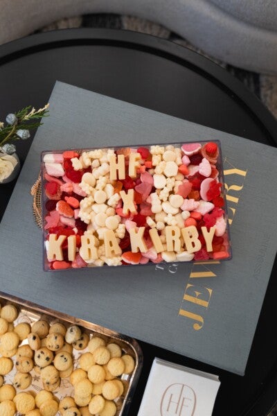 A rectangular clear container filled with assorted red, pink, and white gummy candies and white chocolate pieces spelling out "HF X KIRA KIRBY" on top, placed on a large gray book with gold lettering. Nearby, there is a tray of small round cookies and a stack of white napkins with a circular "HF" logo.