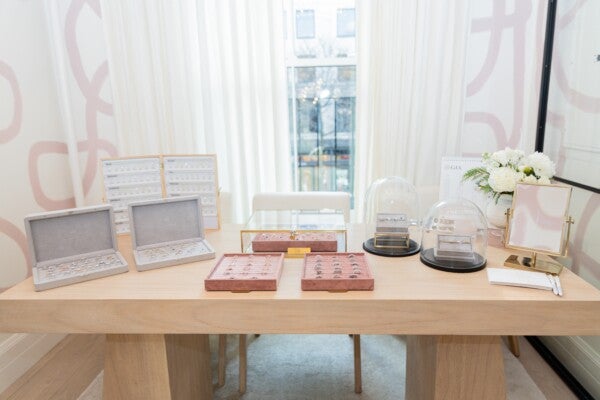 A wooden table displaying various jewelry items, primarily rings, arranged in velvet-lined trays and cases. Two gray cases on the left hold multiple rings, while two pink trays in the center also showcase rings. Behind these, a glass case contains more rings. On the right side, two sets of rings are displayed under glass domes on black bases. A small gold-framed mirror, a notepad with pens, and a white floral arrangement in a vase are also on the right side of the table. The background features white curtains and a window with a view outside.