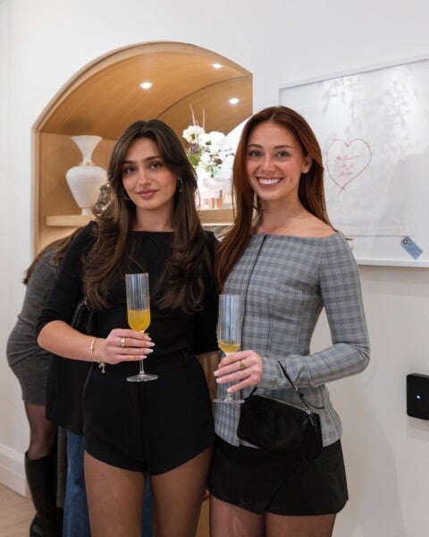 Two young women are standing side by side indoors, each holding a glass of orange-colored drink. The woman on the left has long dark hair, is wearing a black top and black shorts, and accessorized with hoop earrings and bracelets. The woman on the right has long reddish hair, is wearing a gray plaid long-sleeve top and a black skirt, and is holding a small black handbag. Behind them is a wooden arch with decorative items and a framed artwork on the wall.