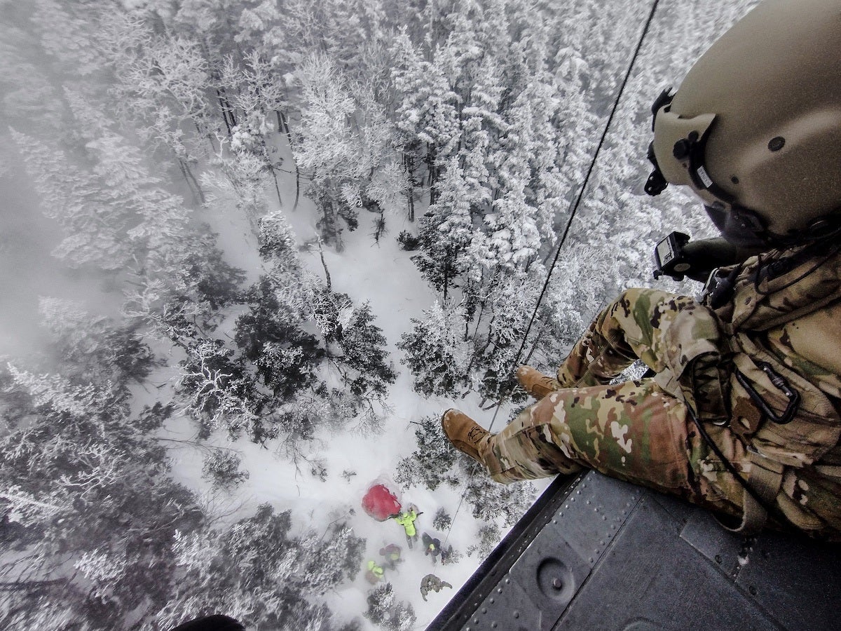 A person in camouflage military gear and helmet is sitting on the edge of a helicopter door, looking down at a snowy forest below. Several people in bright jackets are on the ground near a red object, possibly involved in a rescue or search operation. The forest is densely covered with snow.