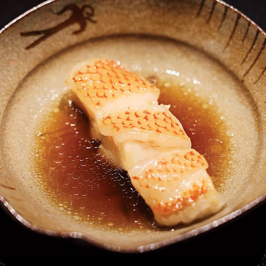 A piece of grilled fish with a golden, slightly charred skin pattern, served in a shallow bowl with a light brown broth. The bowl has a rustic design with a small, dark decorative figure near the rim.