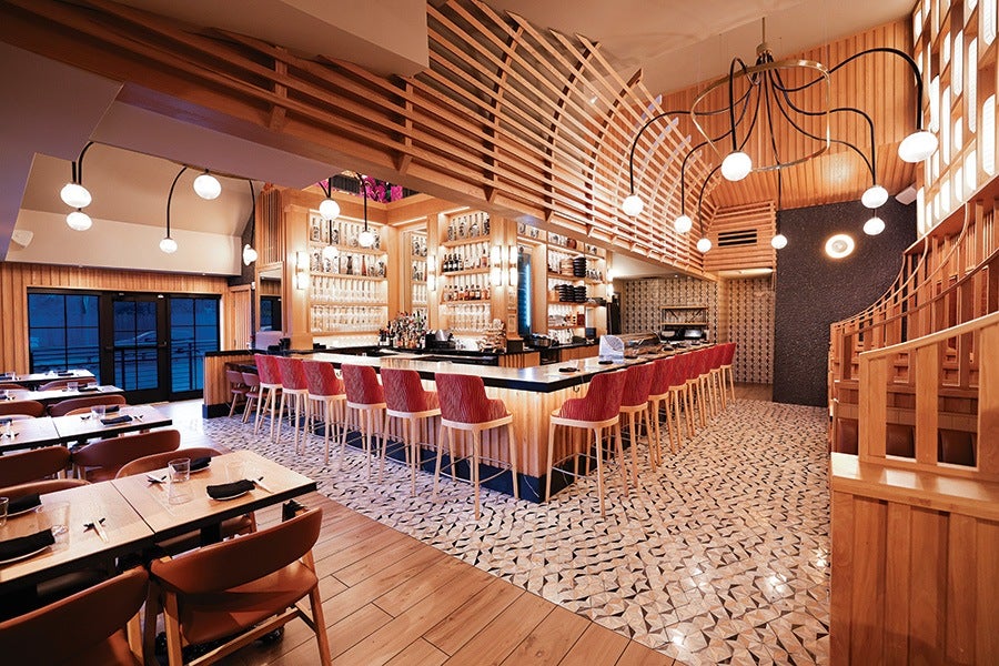 Modern restaurant interior featuring a central bar with red cushioned stools, wooden paneling, and geometric patterned floor tiles. The space is illuminated by multiple round pendant lights with black curved arms, and there are wooden tables and chairs arranged for dining on the left side. The overall design combines warm wood tones with contemporary lighting and decor.