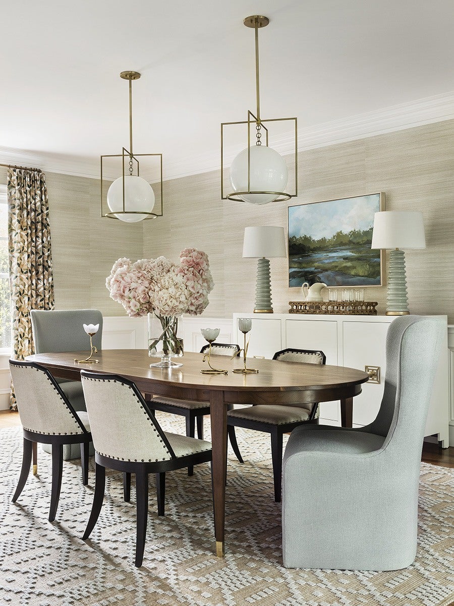 Dining room with a wooden oval table surrounded by six chairs, including two upholstered armchairs in light gray and four beige chairs with dark wooden legs and nailhead trim. A glass vase with pink and white hydrangeas sits on the table, along with three small decorative candle holders. Two modern pendant lights with brass frames and white globes hang above the table. The room features a patterned beige and white rug, a white sideboard with two gray ceramic lamps topped with white shades, and a landscape painting on the wall. Floral-patterned curtains frame a window on the left.