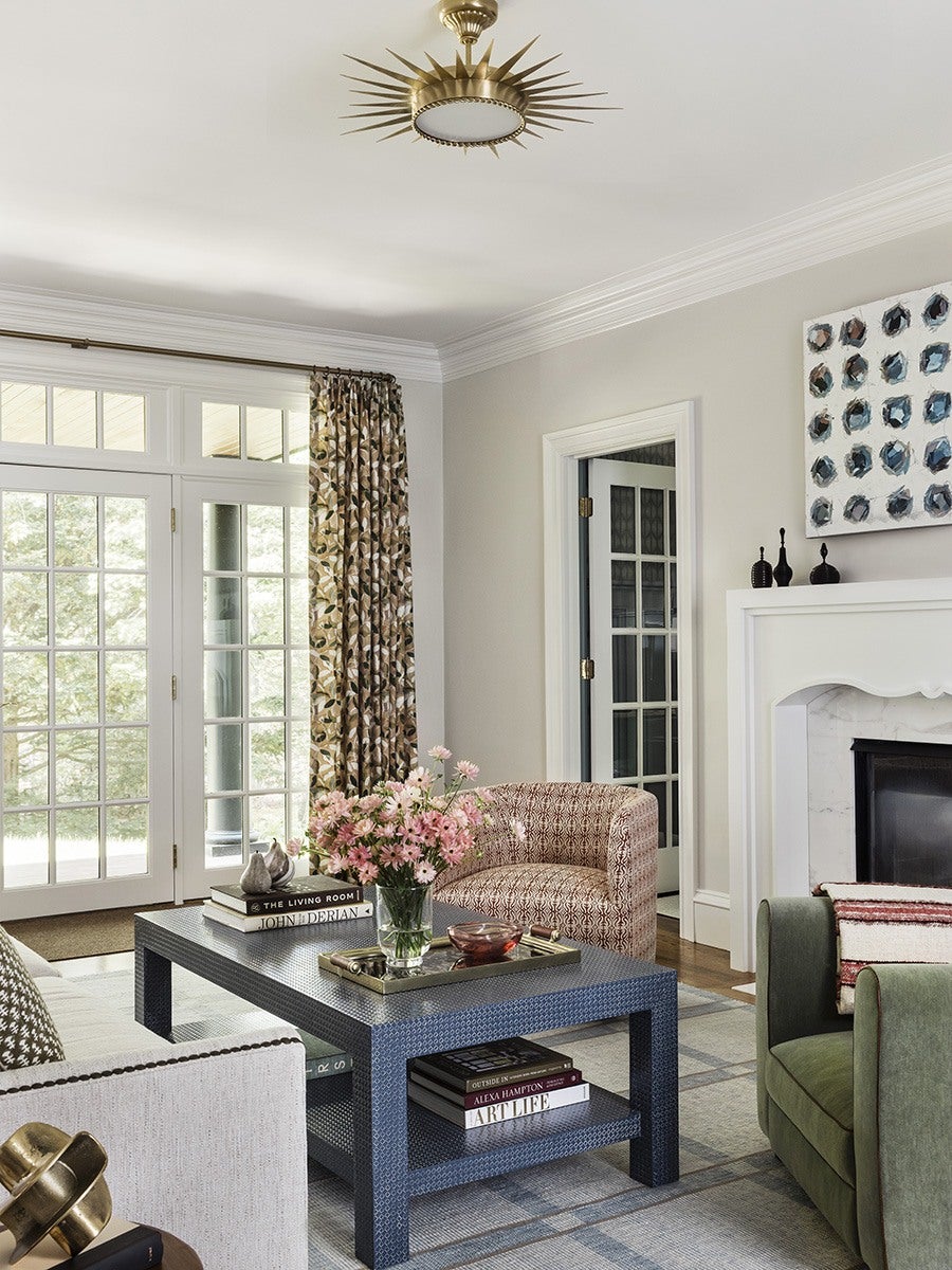 Living room with a blue patterned coffee table holding a vase of pink flowers, books, and decorative items. Surrounding the table are a white sofa with patterned pillows, a pink patterned armchair, and a green armchair with a striped throw blanket. The room features a white fireplace, a large window with floral curtains, and a brass sunburst ceiling light. A modern abstract painting with blue and black dots hangs above the fireplace.