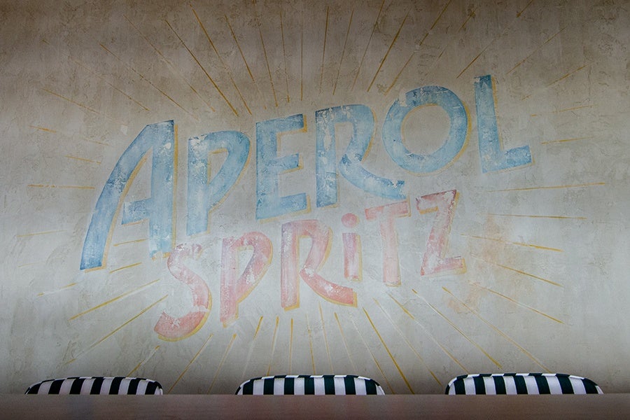 A wall with a faded, vintage-style mural reading "APEROL SPRITZ" in large, colorful letters—blue for "APEROL" and pink for "SPRITZ"—with yellow rays radiating outward. Below the mural are three black and white striped cushions or seats.