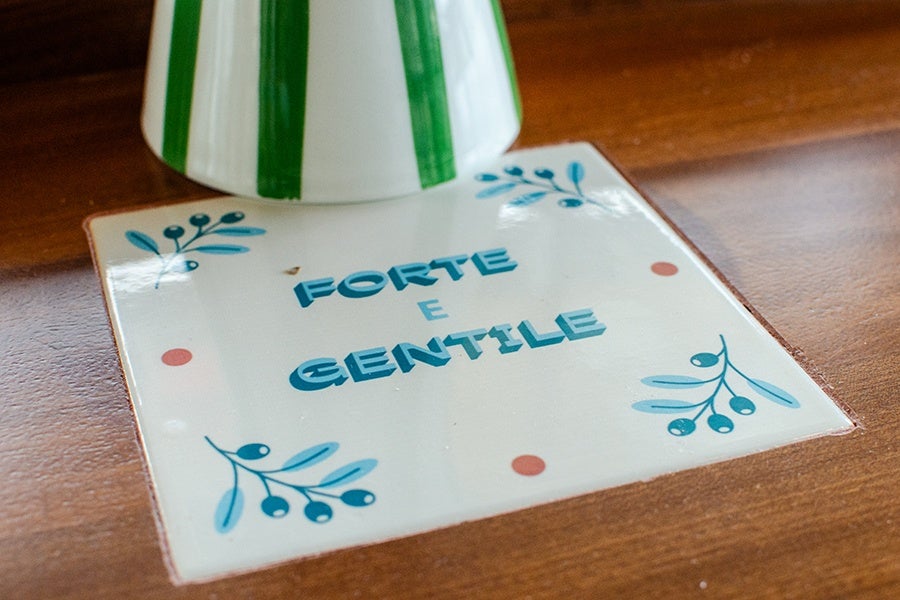 A white ceramic tile set into a wooden surface, decorated with blue leaves and berries in the corners and four small red dots near the edges. The tile has the Italian words "FORTE E GENTILE" written in bold blue letters in the center. A white object with green vertical stripes partially covers the top of the tile.