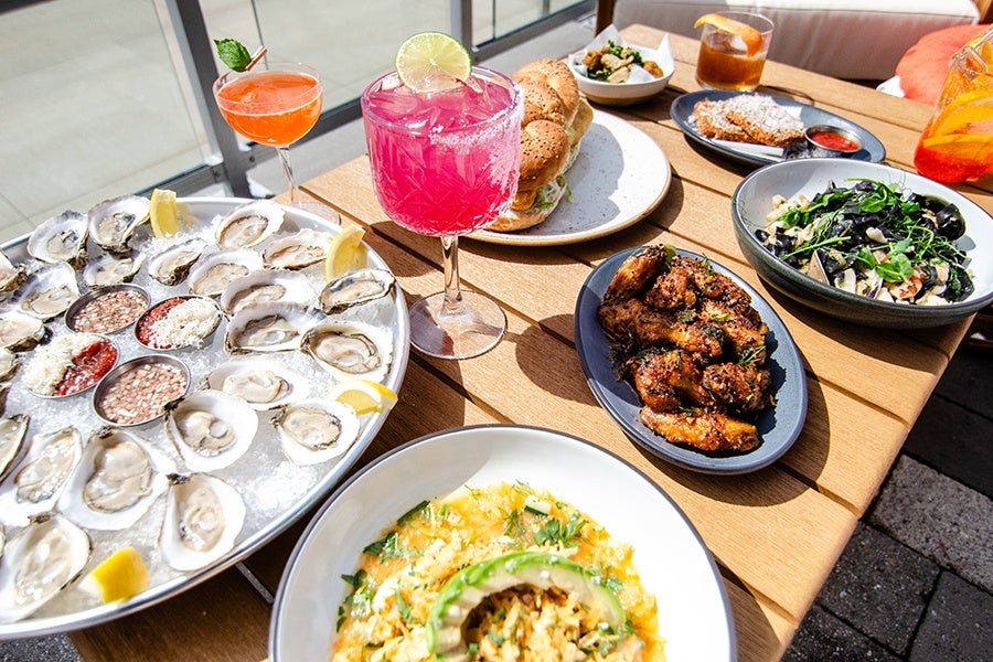 A wooden table set with a variety of dishes and drinks. There is a large platter of fresh oysters on ice with lemon wedges and dipping sauces, a plate of grilled chicken wings, a sandwich on a seeded bun, a bowl of pasta with black squid ink noodles and greens, a bowl of scrambled eggs topped with avocado slices and herbs, and a small bowl of salad. Two colorful cocktails are also present: one bright pink with a lime wheel garnish and another orange cocktail in a coupe glass with a mint leaf garnish. The setting appears to be outdoors with natural light.