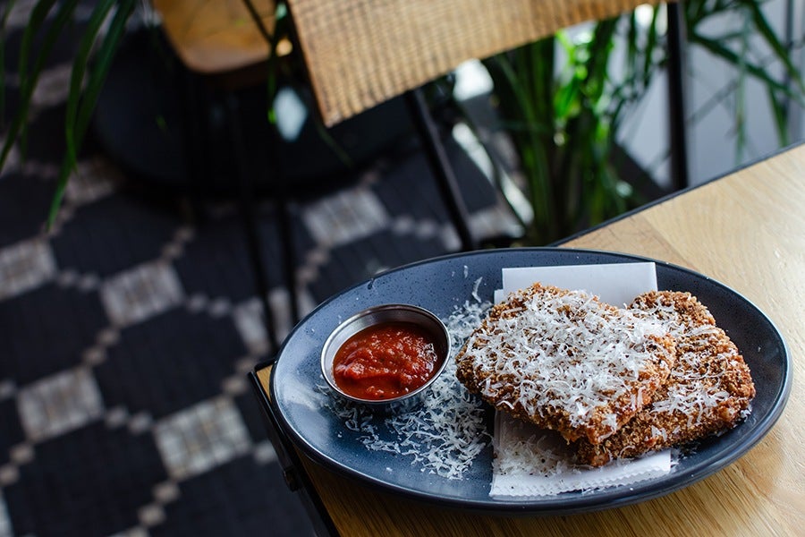 Two breaded and fried square pieces topped with grated cheese on a dark blue oval plate, served with a small metal cup of red marinara sauce, placed on a wooden table.