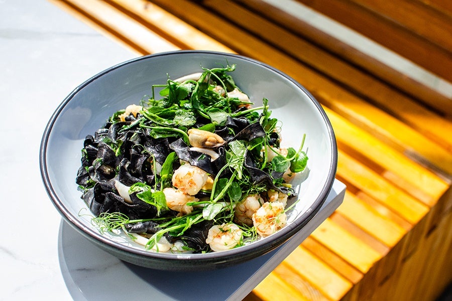 Black squid ink pasta served with shrimp, mussels, and fresh green herbs in a white bowl with a gray rim, placed on a white surface near a wooden bench.