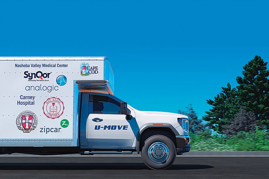 White U-MOVE moving truck with logos of various organizations including Nashoba Valley Medical Center, SynQor, analogic, Carney Hospital, Cape Cod, Bay State College, Eastern Nazarene College, and zipcar on the side, driving on a road with green trees and bushes in the background under a clear blue sky.
