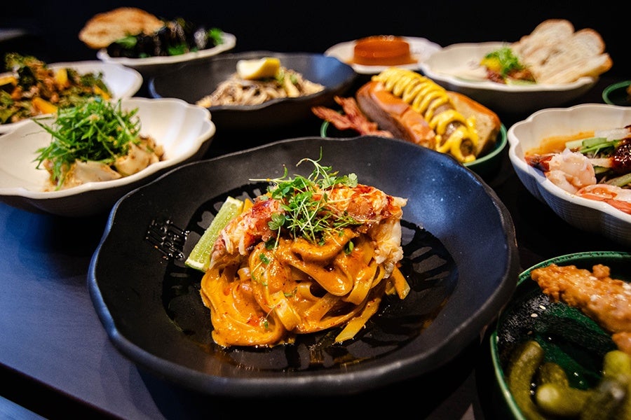 Thai-style small plates are spread across a table, including sausage in a bun, dumplings, noodles in a thick orange sauce, and more.