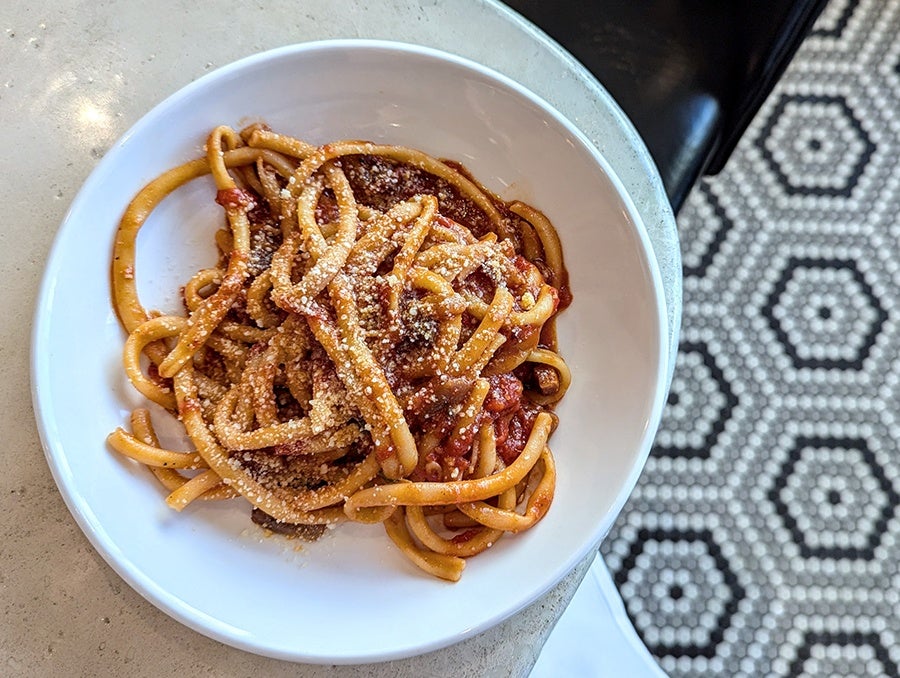 A plate of thick bucatini with tomato sauce and bits of meat sits on a white counter with a black and white tiled floor in the background.