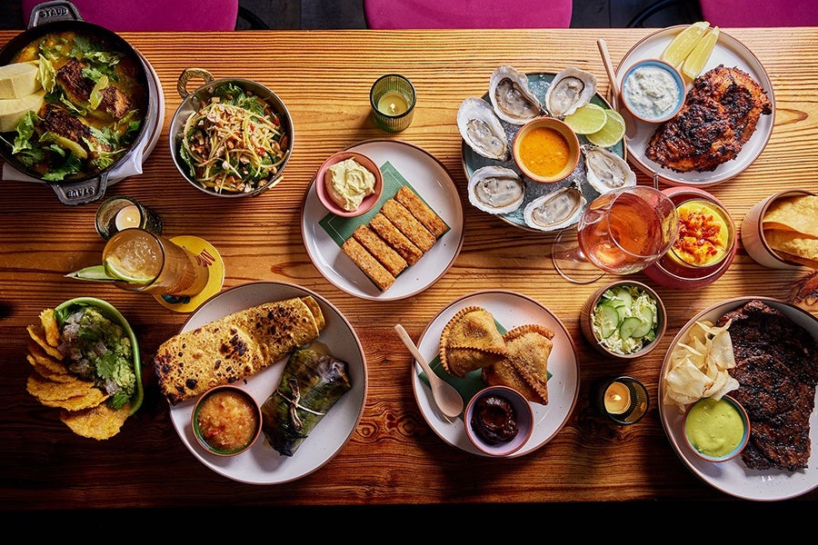 Overhead view of a wooden table covered with food, including samosas, a mango salad, grilled chicken thighs, and more.