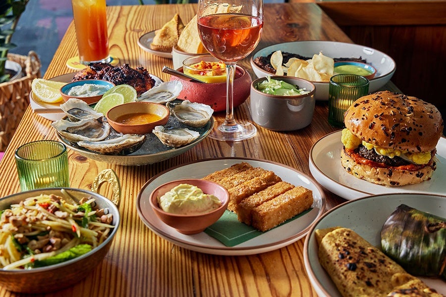 A wooden table is covered with food, including chickpea fries, a cauliflower-topped lamb burger, oysters, and more.