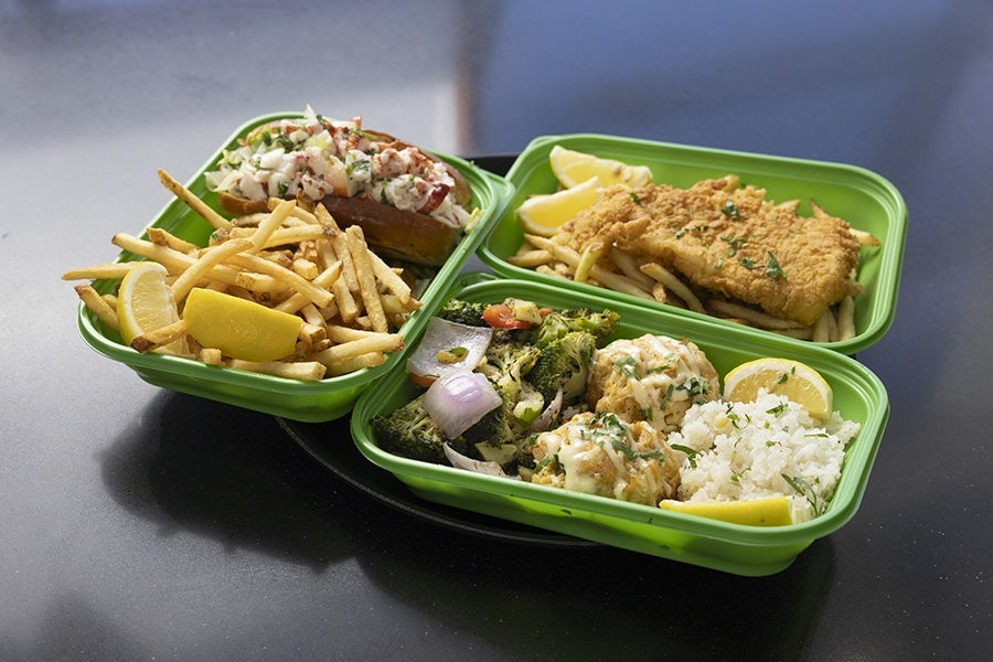 Three green takeout containers with different meals: one with a lobster roll sandwich and French fries garnished with lemon wedges; another with a breaded fried fish fillet on French fries with lemon wedges; and the third with two crab cakes topped with sauce, steamed broccoli and onions, white rice, and lemon wedges.