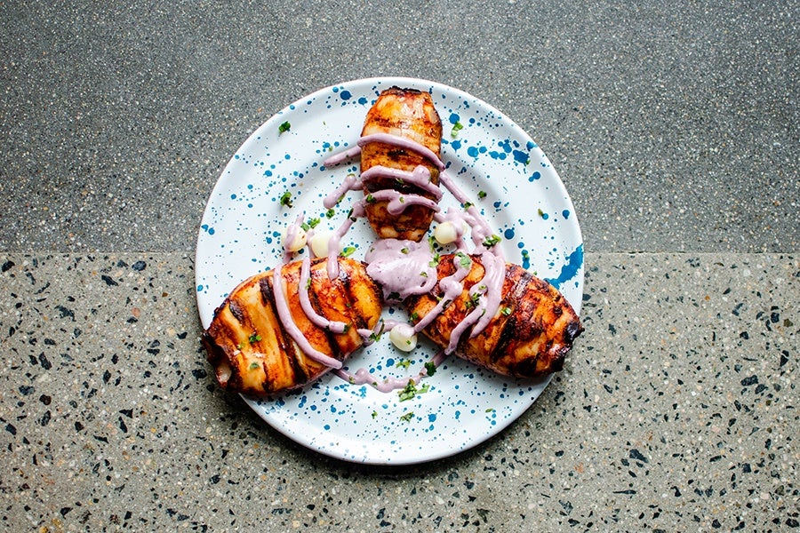 Three grilled pieces of squid are arranged on a white plate with blue splatter patterns. The fish is drizzled with a light purple sauce and garnished with small green herbs and a few dollops of a creamy white sauce. The plate is set on a speckled, textured surface.